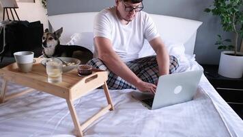 man sitting on the bed using laptop working or playing games and playing with dog video