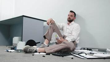 A businessman in a trashed office sits on the floor. Stress, chaos and business man with headache due to paperwork, reports. Concept of burnout, emotional crisis video