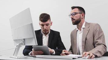 Two business men discuss an online project or presentation at a computer with a tablet. Focused coworkers work in pairs at a computer using apps video