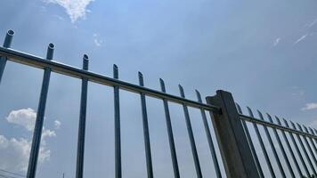 Security Fence Under Blue Sky A close up view of a sturdy metal fence against a vibrant blue sky dotted with fluffy white clouds. The image evokes feelings of security protection and peace of mind. photo