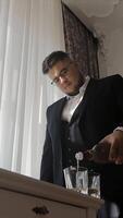 Man in a Suit Pouring a Drink by a Sunlit Window. A man wearing a formal black suit is seen pouring a drink into glasses by a window with white curtains. video