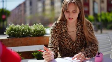 Focused young professional working remotely, writing notes in sunlit outdoor cafe, embodying modern urban lifestyle with creative concentration and relaxed demeanor video