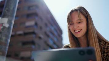Blonde professional using digital tablet while working remotely outdoors, enjoying sunlight and smiling during connection on bright urban street video