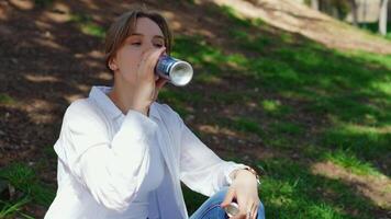 Young woman resting on grassy park lawn, drinking refreshing blue beverage can while enjoying peaceful summer moment, basking in sunlight and surrounding nature video