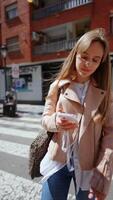 jovem mulher caminhando em cidade pedestre cruzando, navegação Smartphone enquanto explorando urbano ambiente, vestindo na moda Primavera Jaqueta e ficando conectado para digital mundo video