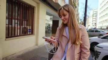 Stylish young blonde woman walking down urban sidewalk, wearing light pink leather jacket and browsing smartphone with confident, casual demeanor during sunny day video
