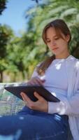 Young woman working remotely on tablet while sitting on park bench, surrounded by green trees and dappled sunlight, embodying digital nomad lifestyle with peaceful concentration video