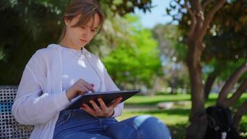 Focused female working on digital tablet while seated on wooden bench, surrounded by verdant park landscape with soft sunlight filtering through trees video