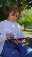 Young woman enjoying leisurely moment in park, working remotely on tablet while sitting on bench, interacting with touchscreen and adjusting hair in relaxed outdoor setting video