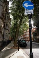 Sunlight shines through trees along a city street, with a road sign indicating a left turn for cycles. The urban scene features a quiet street with a parked vehicle photo