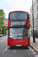 Front view of a red London double decker bus, showcasing the sharp design and clean lines. The surrounding urban landscape and buildings reflect in the bus windows. photo