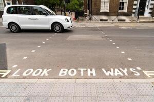A street in London with the phrase LOOK BOTH WAYS painted on the road, indicating the pedestrian crossing. Traffic lights are visible, showing a red signal, and classic London style buildings f photo