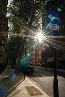 Sunlight shines through trees along a city street, with a road sign indicating a left turn for cycles. The urban scene features a quiet street with a parked vehicle a photo