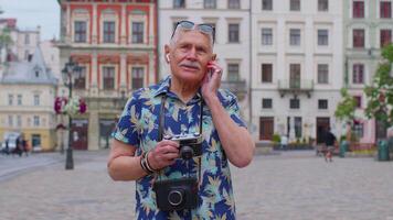 Senior man tourist with retro photo camera, smiling, listening music earphones, dancing on street video