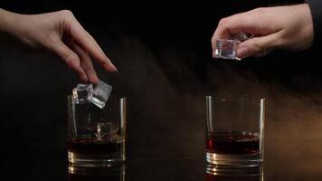 Barmans dropping ice cubes into drinking glasses with whiskey, cognac, brandy on black background video