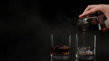 Barman pouring whiskey, cognac, brandy from bottle into glasses with ice cubes on black background video