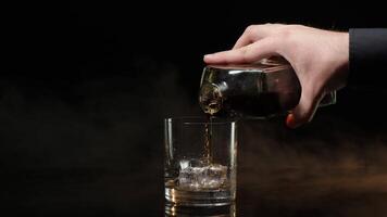 Barman pouring whiskey, cognac, brandy from bottle into glass with ice cubes on black background video