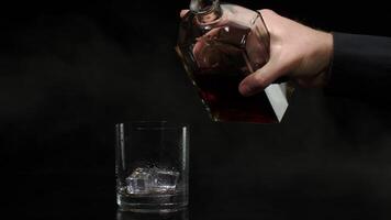 Barman pouring whiskey, cognac, brandy from bottle into glass with ice cubes on black background video