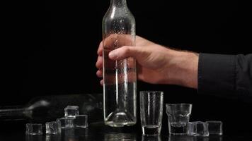 Bartender pouring up two shots of vodka with ice cubes from bottle into glasses on black background video