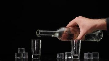Bartender pouring up vodka from bottle into two shots glasses with ice cubes on black background video