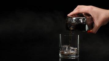 Barman pour whiskey cognac brandy from bottle into drinking glass with ice cubes on dark background video
