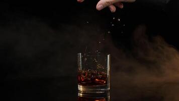 Barman dropping ice cubes into drinking glass with golden whiskey cognac brandy on black background video
