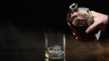 Barman pour whiskey cognac brandy from bottle into drinking glass with ice cubes on dark background video