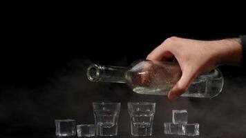 Bartender pouring up two shots of vodka with ice cubes from bottle into glasses on black background video