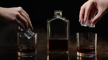 Two hands with glasses of cognac whiskey with ice cubes making cheers toast on black background video