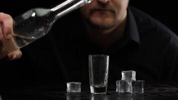 Bartender pouring up frozen vodka from bottle into shot glass with ice cubes on black background video