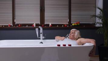 Active senior woman lying in warm bath with bubbles, drinking champagne after hard working day video