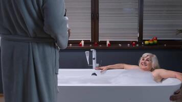 Senior grandmother is taking foamy bath with candles. Grandfather giving glass with champagne video