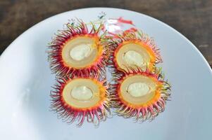 Fresh halved rambutans neatly arranged on ceramic plate. Red hairy peel and translucent flesh stand out. Ideal for food blogs, fruit promos, or exotic recipe visuals. photo