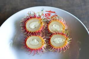 Close up of halved rambutans on a white plate with rustic wood background. Vivid red skin, juicy flesh, and seeds visible. Perfect for tropical food or culinary content. photo