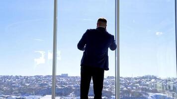 Businessman having a conversation through the phone. Young man in elegant suit standing near the large office window and talking the phone. Rear view. video