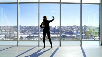 Girl is doing selfie in office. Young woman making photos of herself on a mobile phone while standing near the large office window in a sunny day. Rear view. video