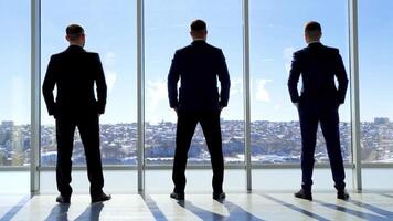 Businessmen looking out of large office window. Back full-length portraits of three entrepreneurs in costumes standing by the window in a office at daytime. Rear view. video
