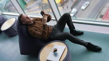 Man rests in a soft chair with a phone. Young man sitting near the window with a city transport background and using his mobile phone. Cup of coffee on small table. Top view. video