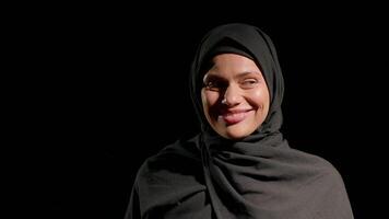 woman in black hijab smiles gently showcasing her warmth and confidence. smells a rose, setting is bright and contrasts dark background emphasizing her expression video