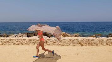 Carefree young female running along sandy shoreline, waving colorful pareu scarf while enjoying summer vacation freedom near ocean waters video