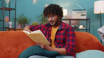 Indian man reading interesting book turning pages smiling enjoying literature taking a rest on sofa video