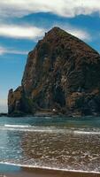 A massive rock formation rises from the ocean on a tranquil beach under a partly cloudy sky. video