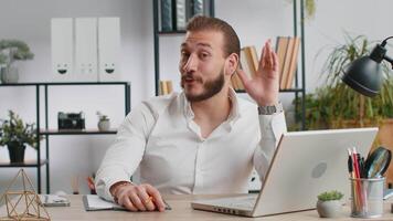 Businessman working on laptop smiling friendly at camera and waving hands gesturing hello at office video