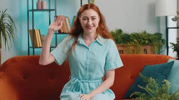 Pretty woman smiling friendly at camera, waving hands gesturing hello, hi, greeting at home on sofa video