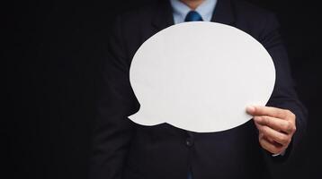 Close-up of a businessman in a suit holding a blank white speech bubble while standing on a black background. Space for text. photo