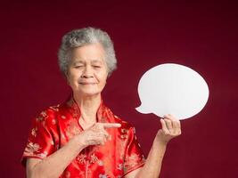 Portrait of a senior woman wearing a traditional cheongsam qipao holding and pointing to a blank speech bubble and smiling while standing on a red background. photo