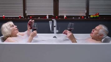 Senior couple man and woman lying in warm bath with bubbles, enjoying relaxation, drinking red wine video