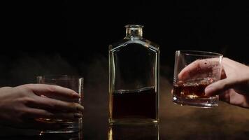 Two hands with glasses of cognac whiskey with ice cubes making cheers toast on black background video