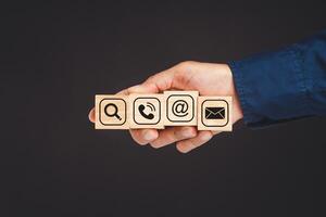 Close-up of a hand businesspeople holding wooden cubes with symbols of a magnifying glass, mobile, address, and email against a blue background. photo