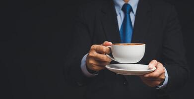Close-up of a businessman in a suit is holding a cup of coffee while standing on a black background. photo